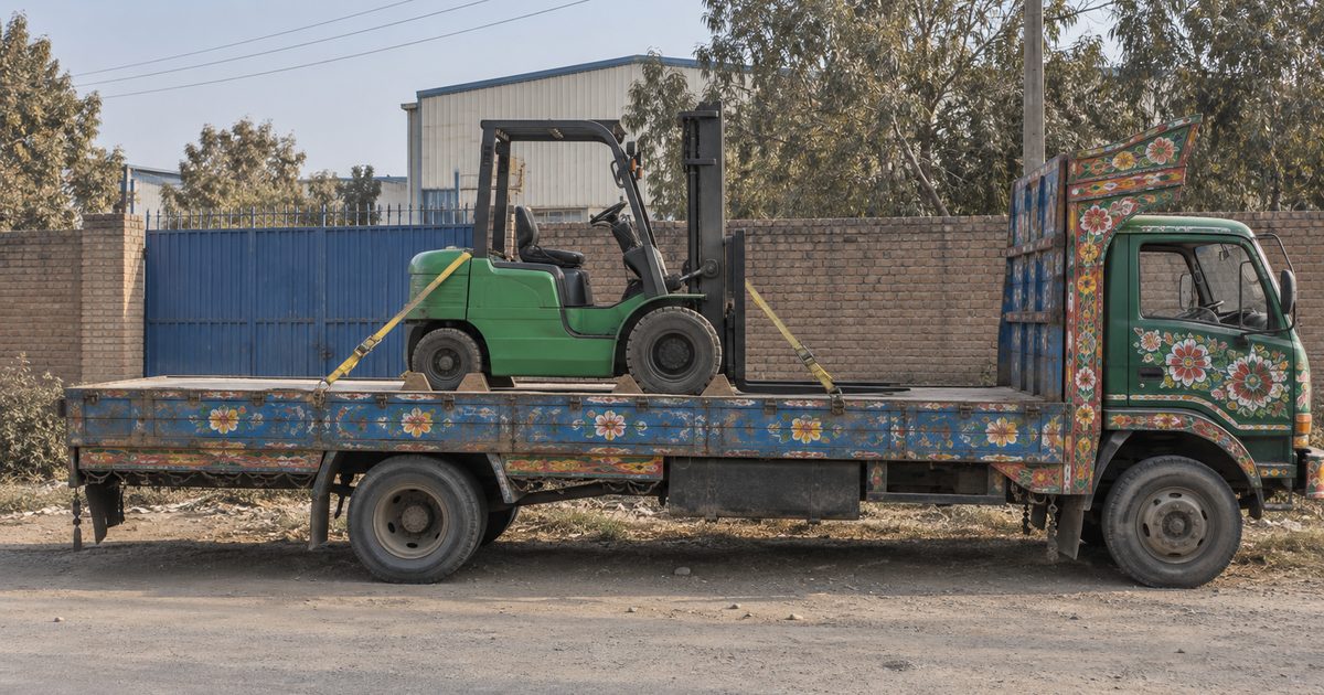 Green forklift strapped and chocked on the bed of a painted goods truck outside a factory