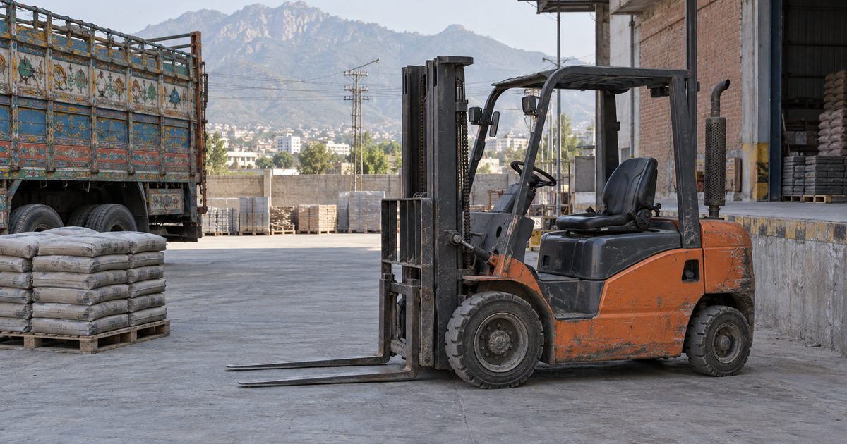 Worn orange forklift parked in a concrete yard beside cement bags and a painted truck